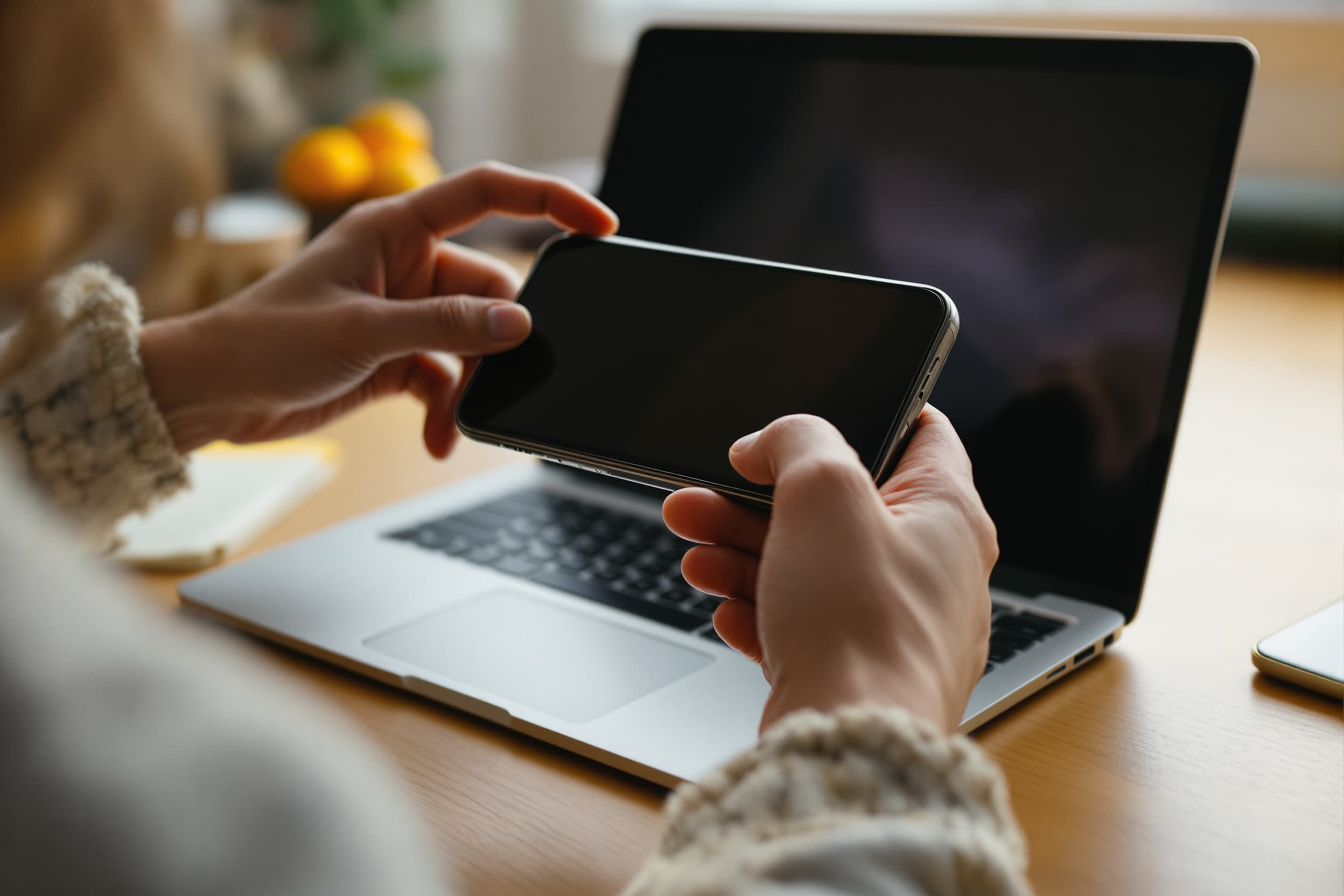 hands holding an iPhone over a laptop at a warm desk